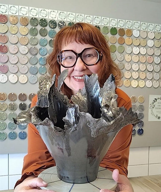 Anna med stor vas, toppigt avslut. Vasen är brun/grå ser nästan metallisk ut. Bakom på väggen är det massa glasyrprover.